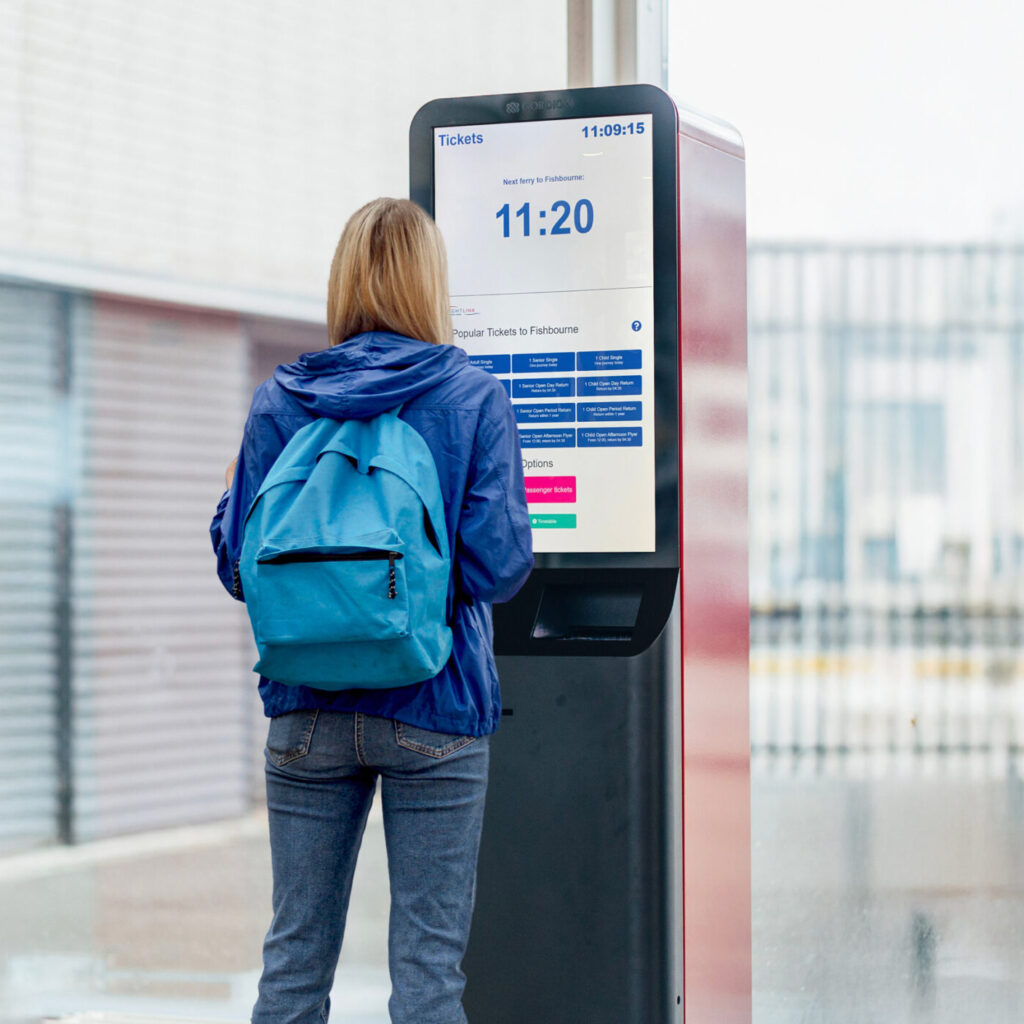 Person med blå ryggsekk bruker en selvbetjent kiosk for billetter utendørs.