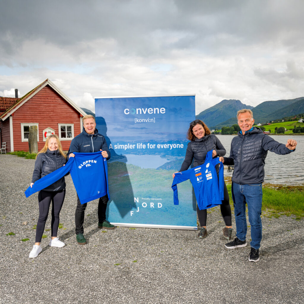 Bilde av fire smilende personer som står foran et banner med teksten "Convene - A simpler life for everyone". To av personene holder opp blå gensere med "Gloppen FIL" skrevet på ryggen. I bakgrunnen er natur med fjell, vann og et rødt hus.